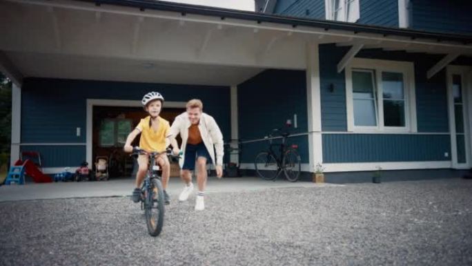 快乐的父亲带儿子在他们家前院的草坪上骑自行车。小男孩戴着头盔，骑自行车保持平衡。骄傲的爸爸为他的成功