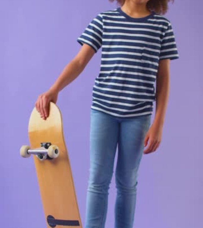 微笑男孩站在滑板上看着紫色工作室背景下的相机的垂直视频-慢动作拍摄