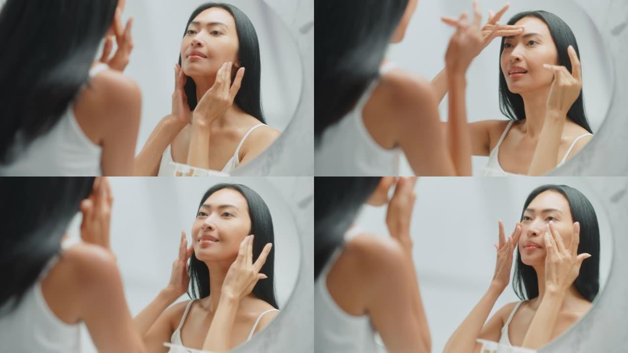 美丽的亚洲女人她完美柔软的面部皮肤，使用先进的有机面霜，微笑在镜子里。快乐的女性享受她早上的美容习惯