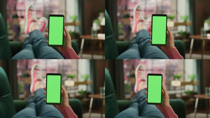 带有绿屏模拟显示的智能手机上的女性手滚动提要。女性在家里的沙发上放松，在移动设备上观看视频和阅读社交