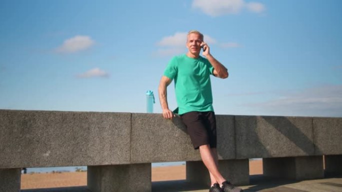 一位金发碧眼的斯堪的纳维亚跑步者正靠在路堤的石栏上，在电话上完成了对话。