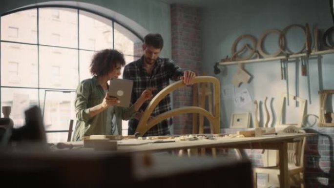 两个才华横溢的小企业主使用平板电脑，并在家具车间讨论新木椅的设计。木匠和一个在阁楼工作室工作的年轻女