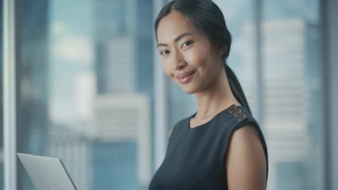 一位穿着时髦的黑色连衣裙的亚洲女商人的美丽肖像，使用笔记本电脑，在城市办公室的窗户旁边摆姿势。自信的