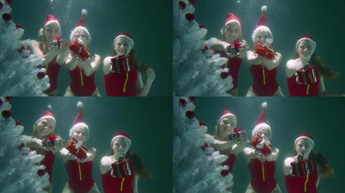 三个穿着红色泳衣，帽子的快乐微笑的仙女妇女在圣诞树水下慢动作中向相机赠送礼物。