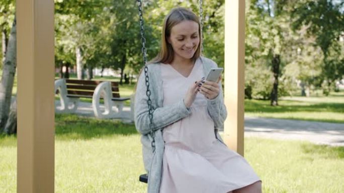 快乐孕妇的慢动作在城市公园的秋千上放松，并使用智能手机微笑