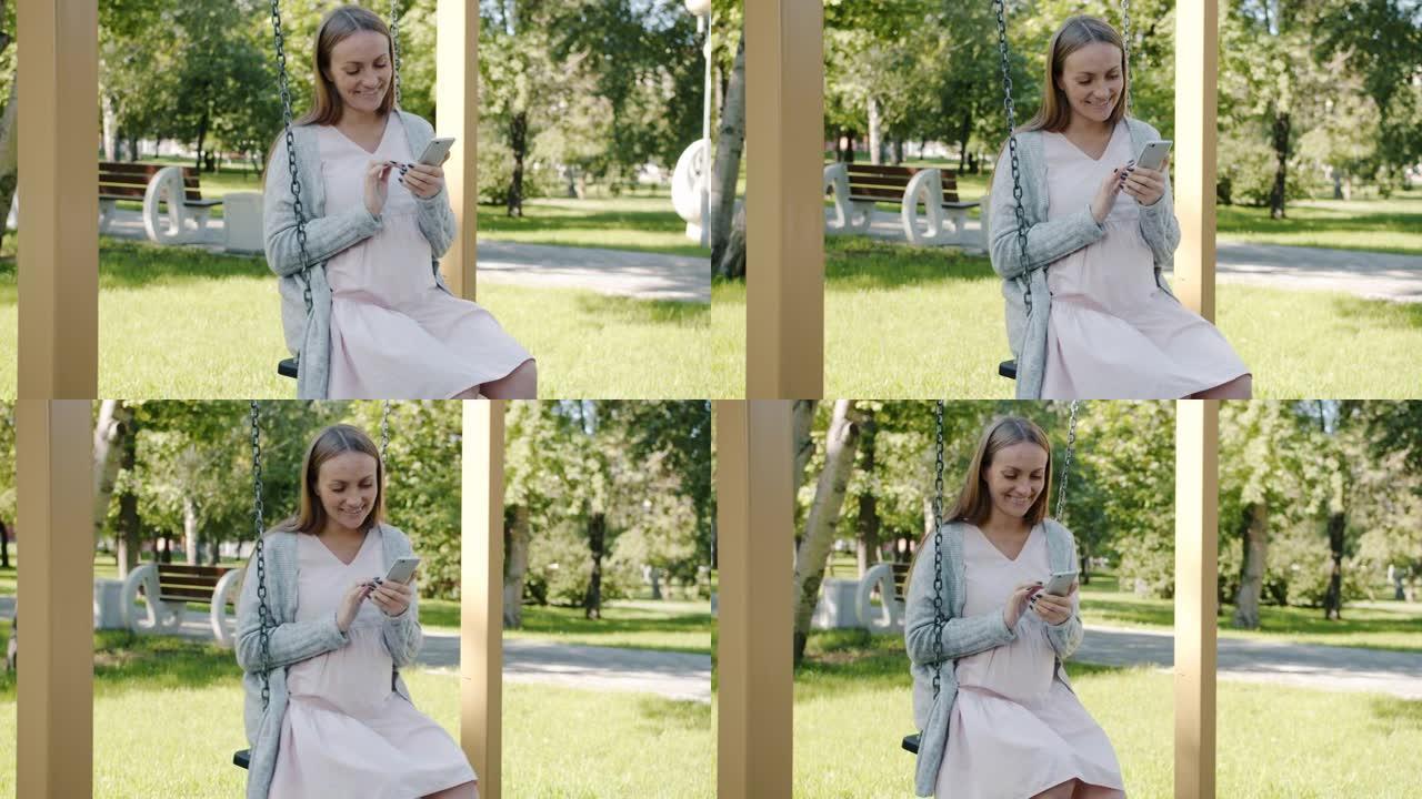 快乐孕妇的慢动作在城市公园的秋千上放松，并使用智能手机微笑