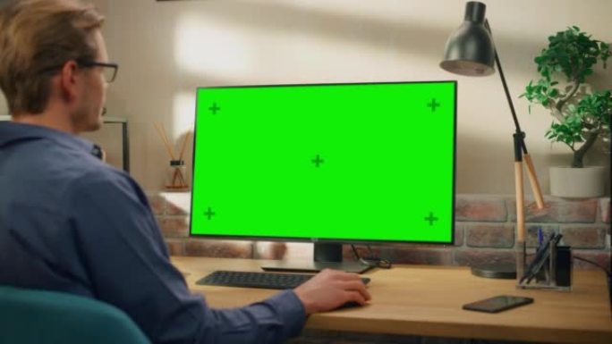 年轻英俊的男人在家工作，在带有绿屏模拟显示的台式计算机上。创意男性查看社交媒体，浏览互联网。明亮阁楼