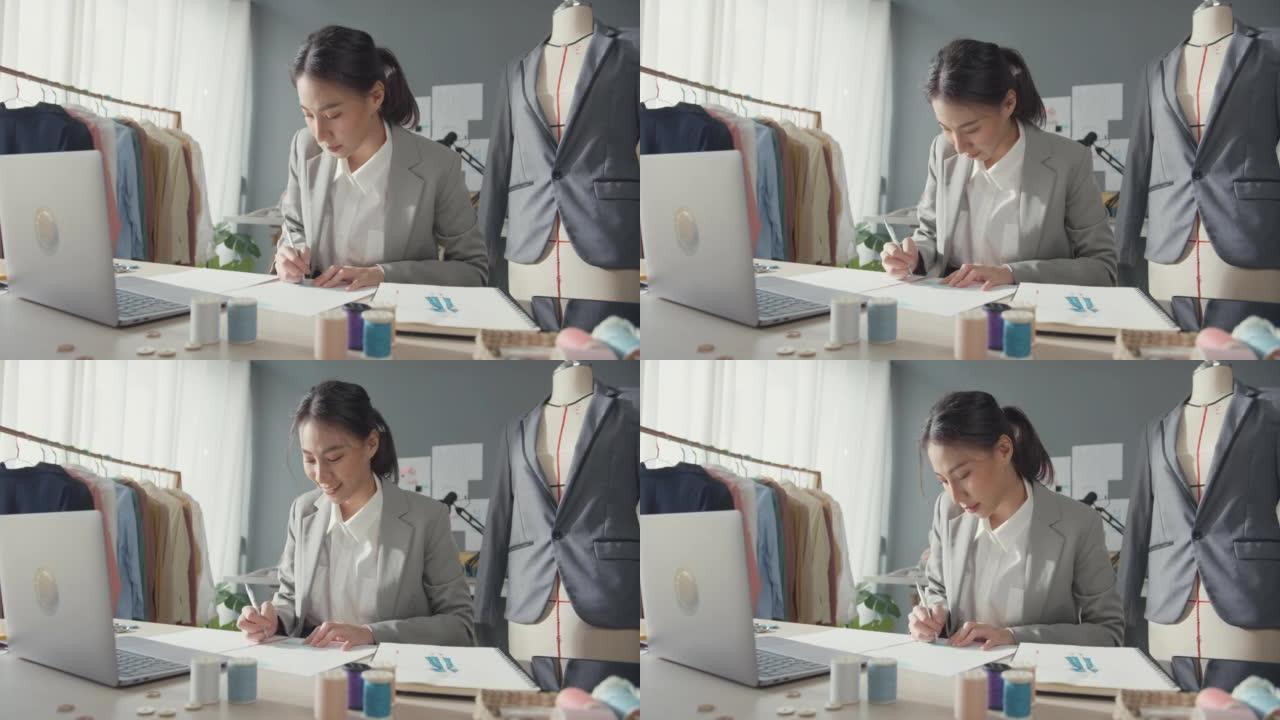专业开朗的亚洲女商人设计师坐在桌子上，用笔记本电脑绘制设计服装时尚工作室工作项目素描本新系列。成功的