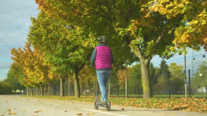 一名老年妇女在秋天的树木旁驾驶电动踏板车的后视图