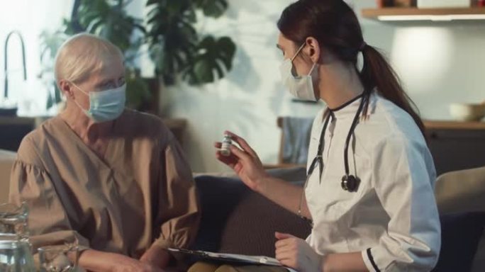 穿着实验室外套的女医生在家访期间与戴着医用口罩的60多岁女性交谈，并提供装有COVID疫苗的烧瓶。