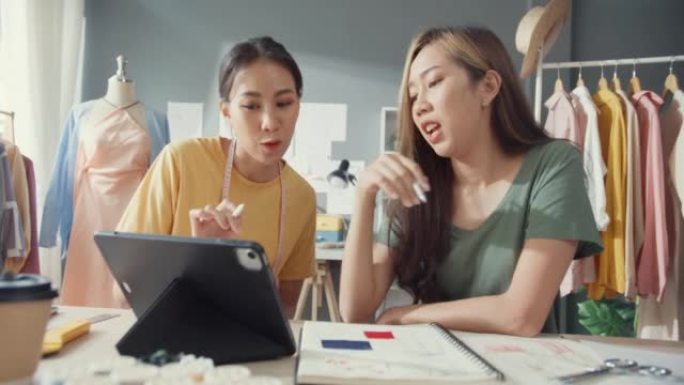 两位专业的亚洲开朗女士时装设计师团队合作，在餐桌上休闲网站使用平板电脑会议讨论最终设计分享想法。女裁