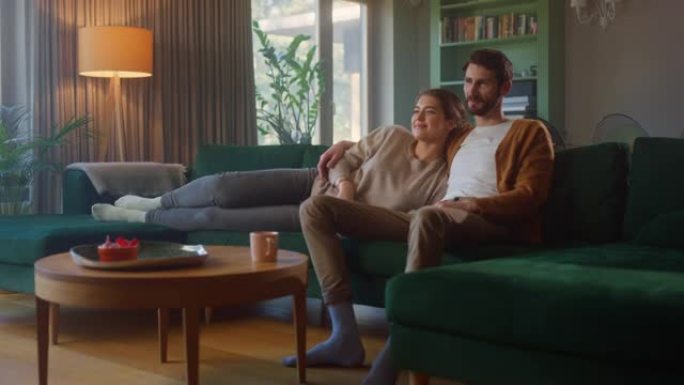 夫妻坐在客厅的沙发上一起看电视。女友和男友拥抱，拥抱，交谈，微笑和观看电视流媒体服务。拥有舒适时尚内