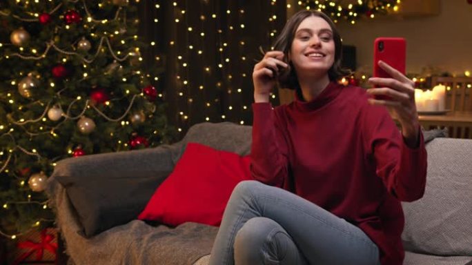 一个穿着红色毛衣和牛仔裤的棕色头发的年轻漂亮女人坐在装饰精美的舒适圣诞节房间里，在她的红色智能手机上
