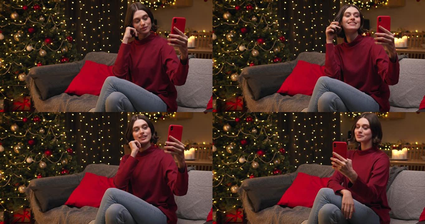 一个穿着红色毛衣和牛仔裤的棕色头发的年轻漂亮女人坐在装饰精美的舒适圣诞节房间里，在她的红色智能手机上