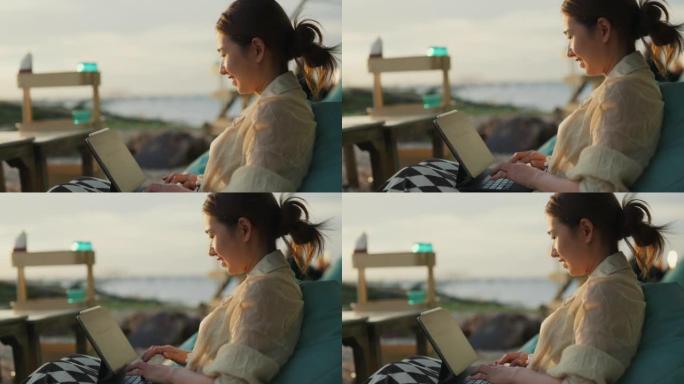 快乐的亚洲女人坐在海边的餐厅时使用数字平板电脑。