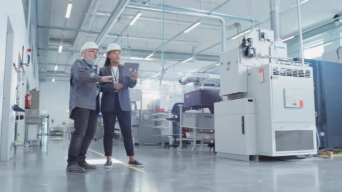 工厂里两名戴着安全帽的重工业员工的肖像。使用平板电脑检查和讨论工业设施。非裔美国工程师和中年技术人员
