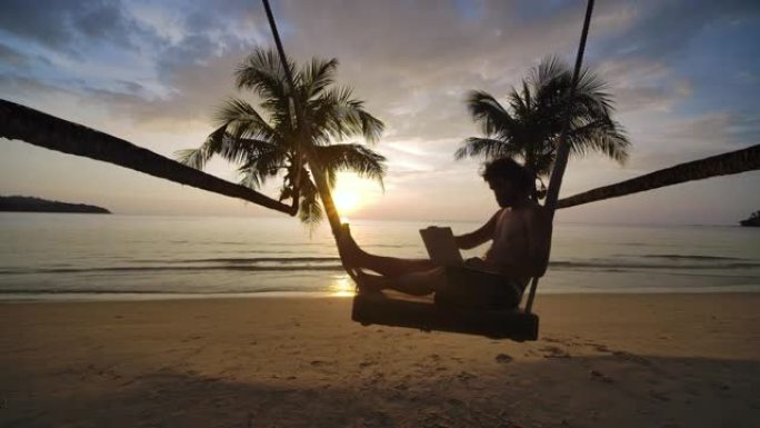暑假旅行的商人自由职业者，坐在笔记本电脑上的旅行者坐在秋千上，在海滩上摇曳，在热带岛屿上慢动作享受夏