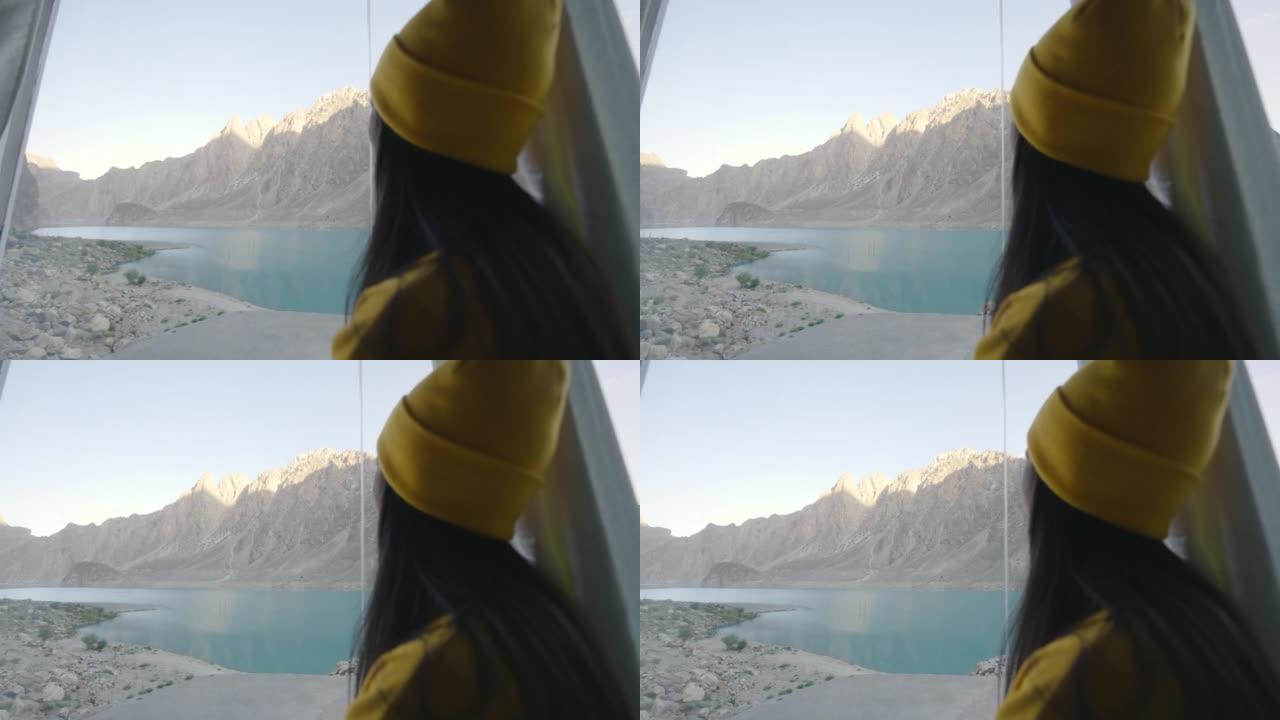 穿着黄色保暖衣服的年轻女子的后视图透过湖边的窗户看。种族多样化的旅行者概念。与Alpha chane