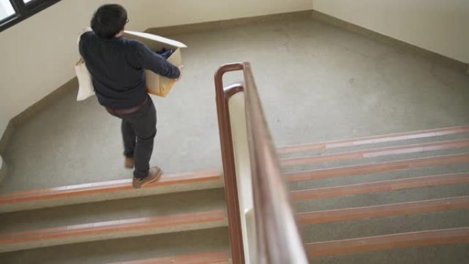 亚洲男子提着一个箱子，里面装着许多办公物品走在楼梯上