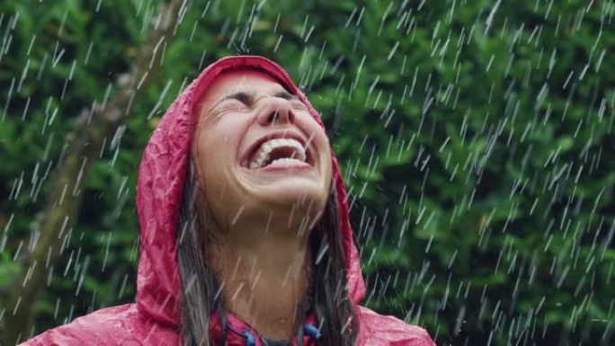 一个穿着红色雨衣的快乐女人的肖像，笑着享受在雨下。女性成年人感激水和自然的祝福，周围是树木。秋乐概念