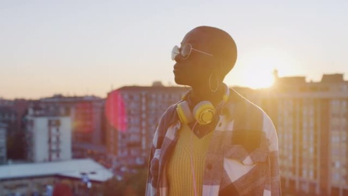 年轻时尚的黑人女性戴着太阳镜和耳机，站在城区的屋顶上。美国黑人女性享受日落和温暖的肖像