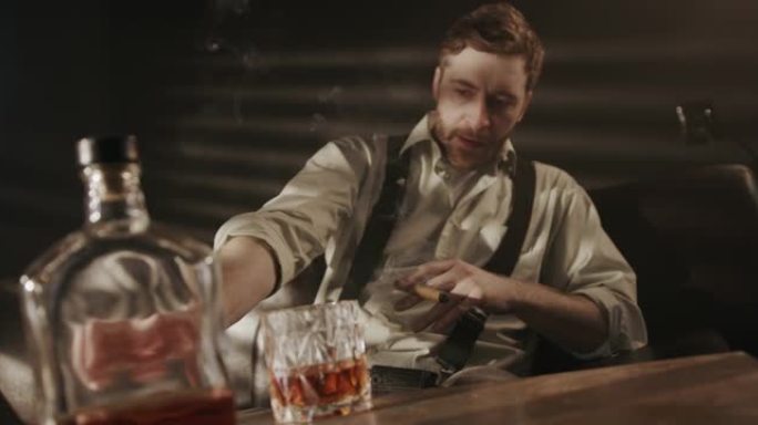 一个穿着衬衫和吊带的喝酒的人点燃雪茄，然后在黑暗的房间里的沙发上抽烟，视频的前景是一瓶威士忌和一杯