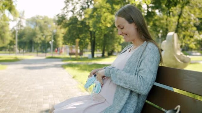 期待母亲玩婴儿鞋的肖像在腹部微笑着等待坐在城市公园长椅上的孩子