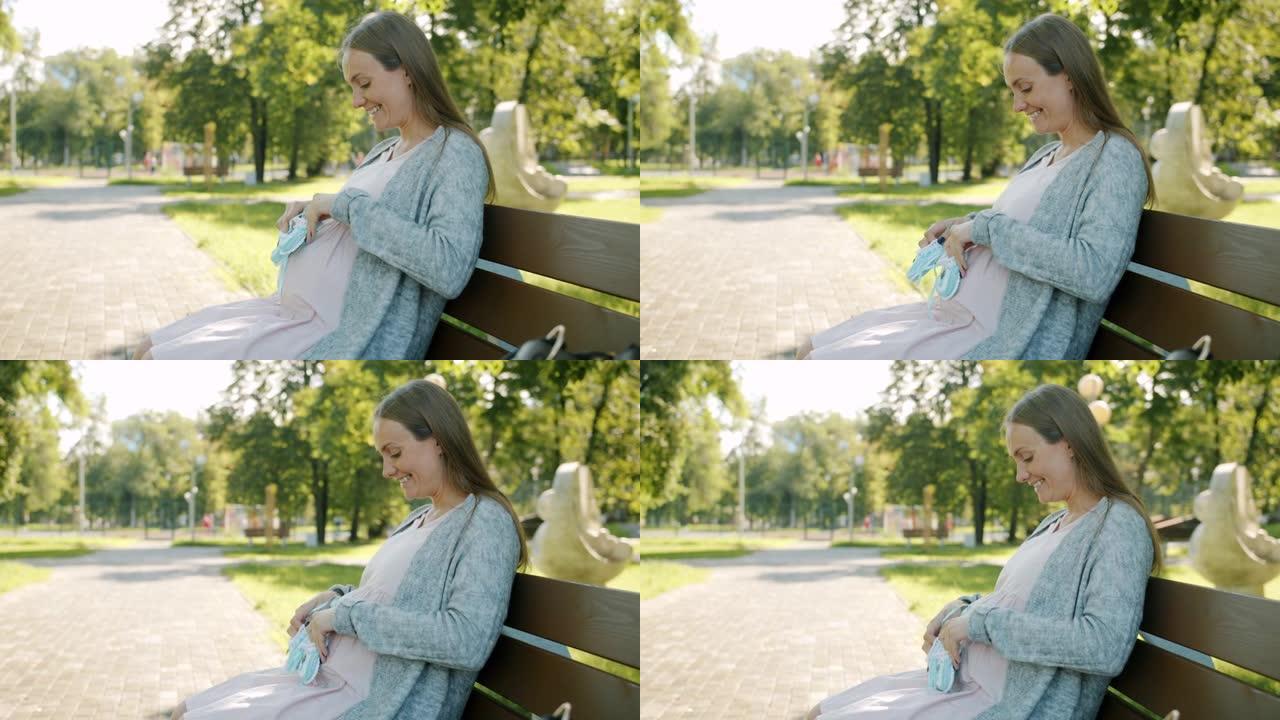 期待母亲玩婴儿鞋的肖像在腹部微笑着等待坐在城市公园长椅上的孩子
