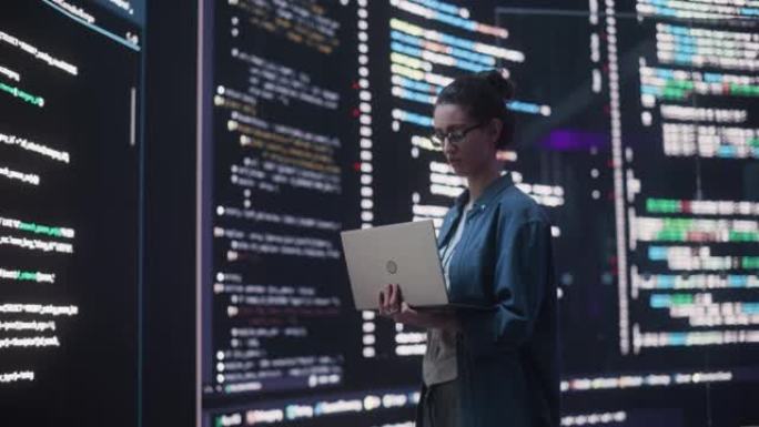 在笔记本电脑上工作的年轻女子的肖像，看着显示后端代码行的大数字屏幕。专业程序员开发大数据接口软件项目
