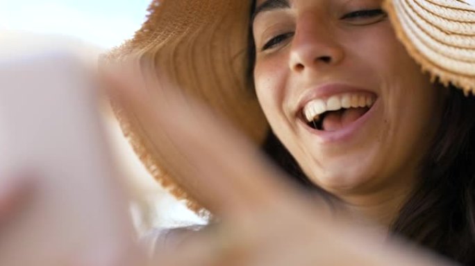 一位戴着草帽的快乐无忧无虑的微笑年轻女性游客在阳光明媚的日子里，正在使用智能手机发送消息，拨打电话或