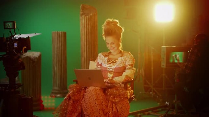 在古装戏剧电影中: 美丽的微笑女演员穿着文艺复兴时期的连衣裙，坐在椅子上，使用背景为绿屏的笔记本电脑
