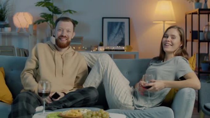 幸福的年轻夫妇看电视的肖像喝着酒杯，在黑暗的房子里微笑