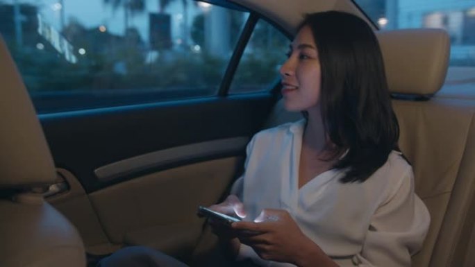 穿着时尚办公服装的亚洲女商人晚上在城市现代城的汽车后座上使用智能手机工作到很晚。