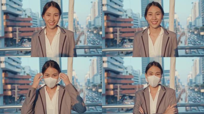 穿着时尚办公服装的亚洲女商人戴着医用口罩，微笑着看着相机，快乐地站在城市现代城市中独自一人。