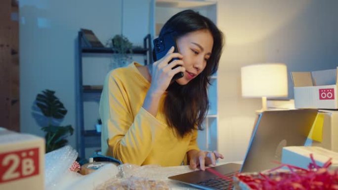 年轻的亚洲女性打电话给智能手机与客户交谈，以检查晚上在家庭办公室的笔记本电脑上确认库存订单。