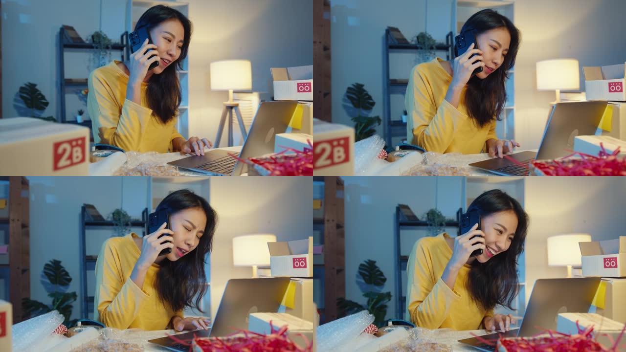 年轻的亚洲女性打电话给智能手机与客户交谈，以检查晚上在家庭办公室的笔记本电脑上确认库存订单。