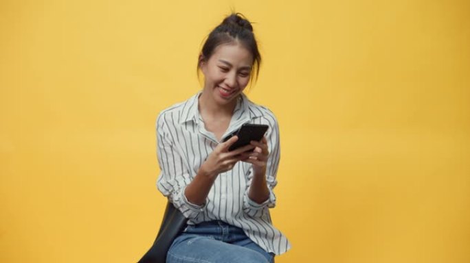 快乐美丽亚洲青年休闲女孩微笑享受打字智能手机信息与朋友站在孤立的黄色空白空间背景。