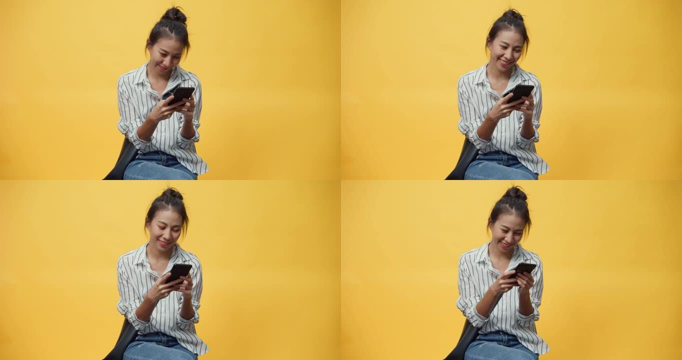 快乐美丽亚洲青年休闲女孩微笑享受打字智能手机信息与朋友站在孤立的黄色空白空间背景。