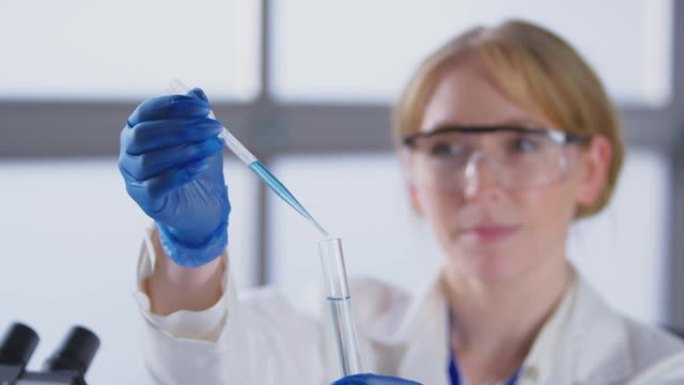穿着白大褂的实验室女工作人员将蓝色液体从移液管中注入试管