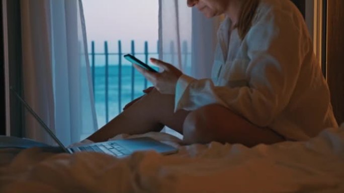 金发碧眼的女人坐在笔记本电脑前的酒店房间的床上，使用智能手机，放下手机，在阳台上散步，欣赏傍晚的景色