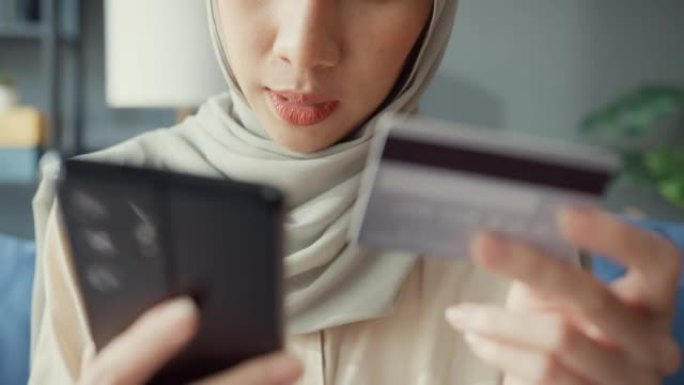 特写迷人的年轻年轻的亚洲伊斯兰穆斯林美女戴着头巾，随意坐在沙发上使用手机在家里舒适的客厅用信用卡享受