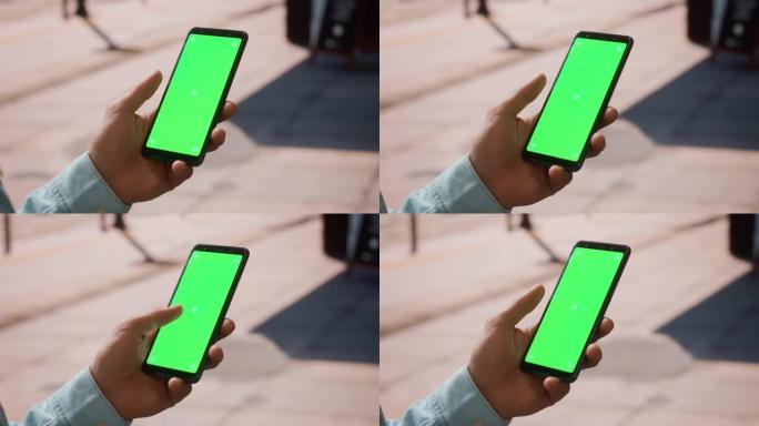 在阳光明媚的日子里，一个年轻人使用带有绿色屏幕色度键模拟显示的智能手机在街道上垂直显示。他正在轻敲屏