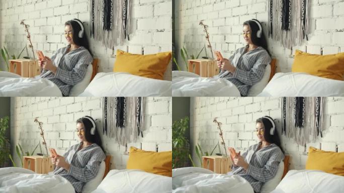 可爱的亚洲女士的肖像，戴着眼罩，在家里的床上戴着智能手机，通过耳机听音乐