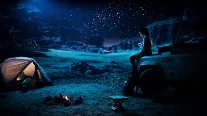 坐着的女性旅行者使用智能手机，在篝火旁在峡谷中露营时凝视着星空。自信的女冒险家在社交媒体互联网上分享