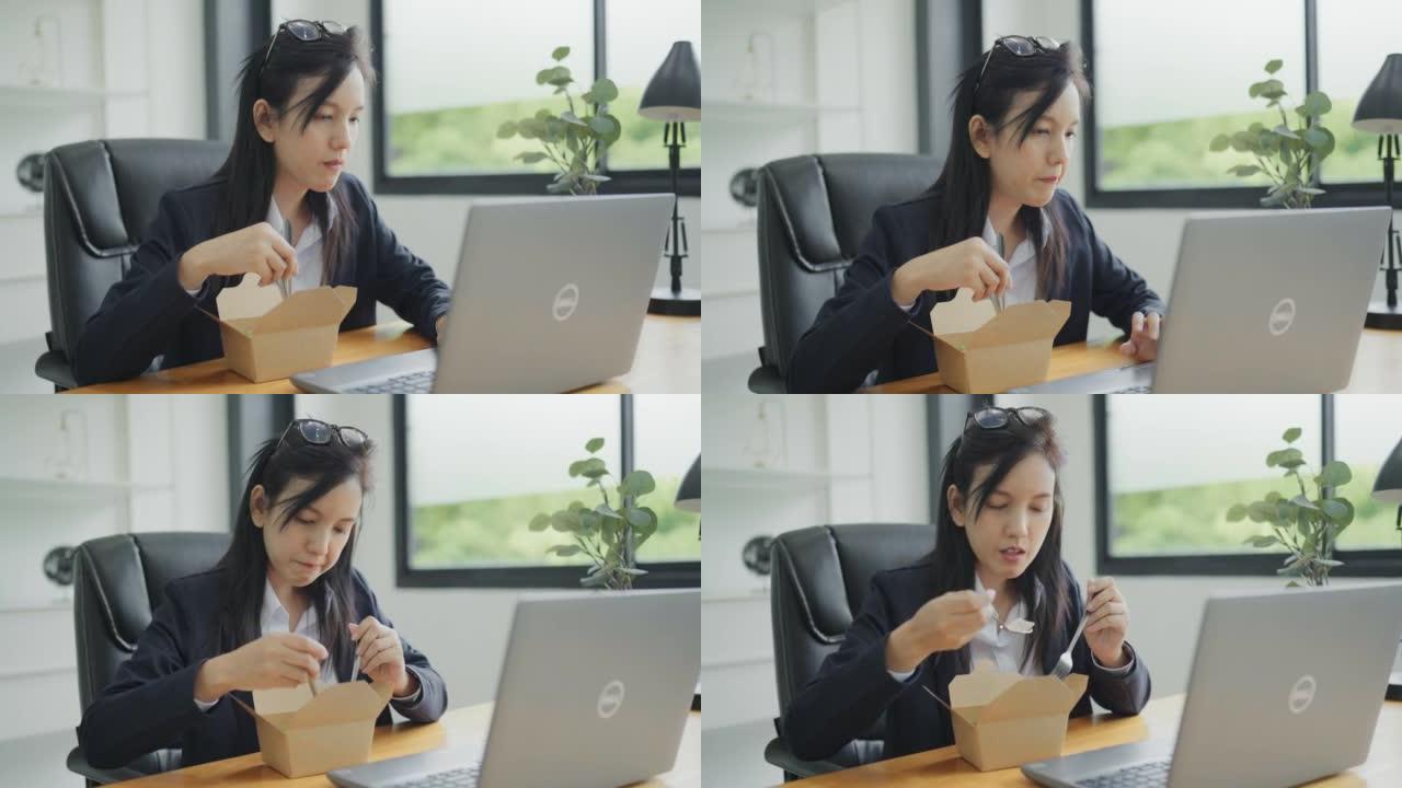 亚洲妇女在笔记本电脑上工作，并在办公室的办公桌工作场所吃沙拉餐