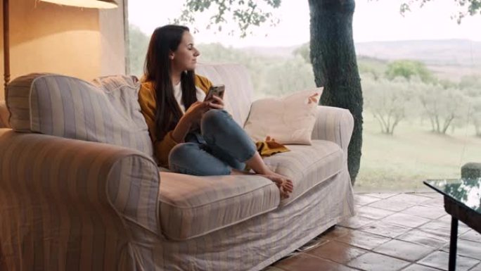 DS年轻女子在托斯卡纳一所房子的露台上的沙发上使用智能手机