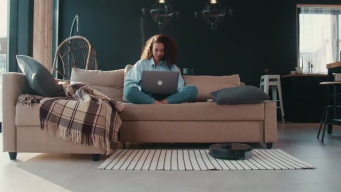 美丽快乐的年轻非裔美国自由职业者妇女在家工作，使用笔记本电脑，机器人清洁扫地。