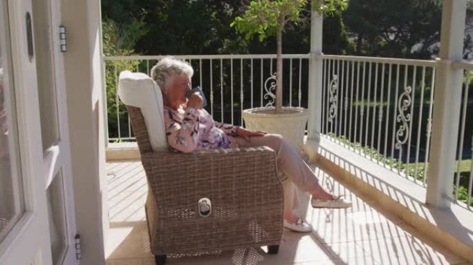 白人高级妇女坐在家里阳台的椅子上喝咖啡