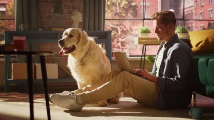 年轻人坐在地板上，在舒适的阁楼客厅里工作或学习笔记本电脑。金毛猎犬狗坐在他旁边，想玩耍并被爱抚。