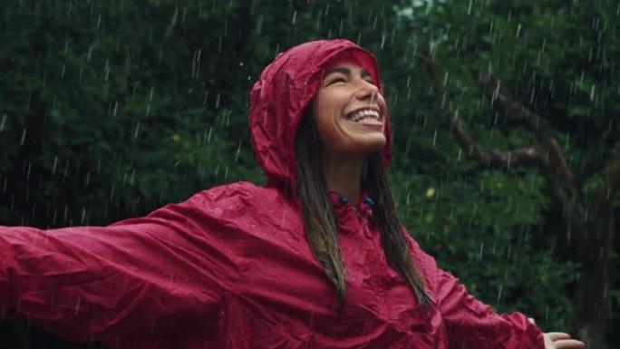 一个穿着红色雨衣的快乐女人的肖像，笑着享受在雨下。女性成年人感激水和自然的祝福，周围是树木。秋乐概念
